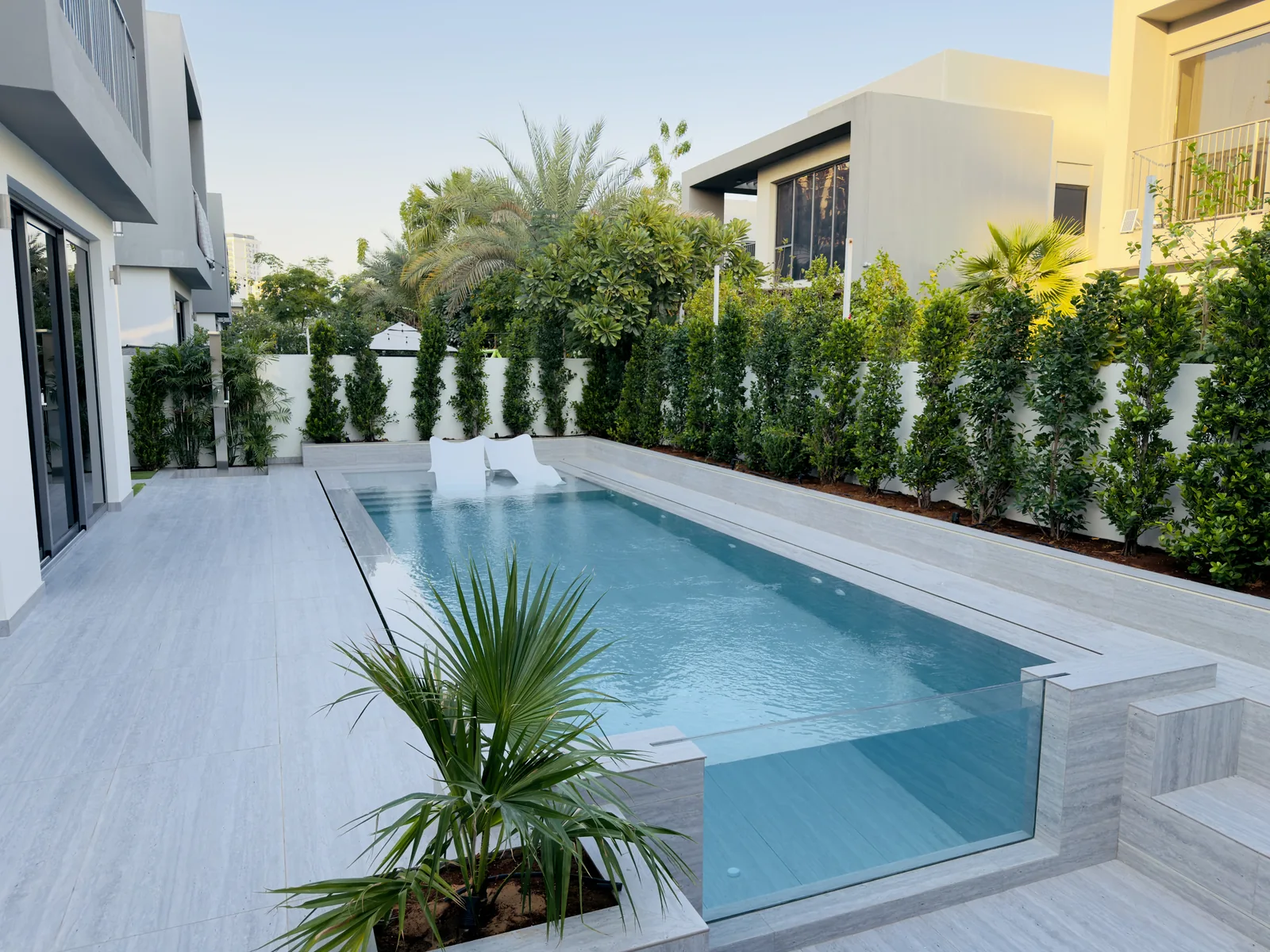 Swimming pool beside a villa with pale stone paving and boundary planting